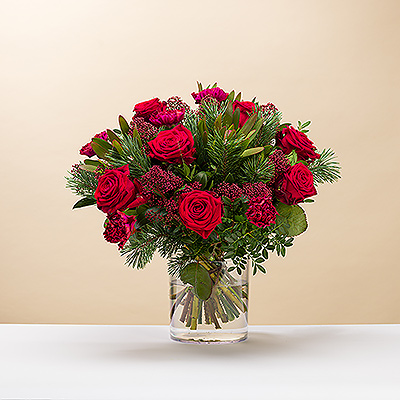 Des voeux de Noël avec un magnifique bouquet rouge et vert. Des roses rouges classiques et d'autres fleurs sont complétées par des branches de pin et de la verdure dans ce bouquet de Noël festif.