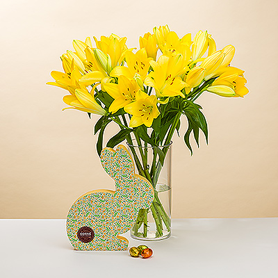 Brille con un ramo de preciosos lirios amarillos frescos y pequeños huevos de chocolate Corné Port-Royal en una encantadora caja de regalo con forma de conejo de Pascua.
